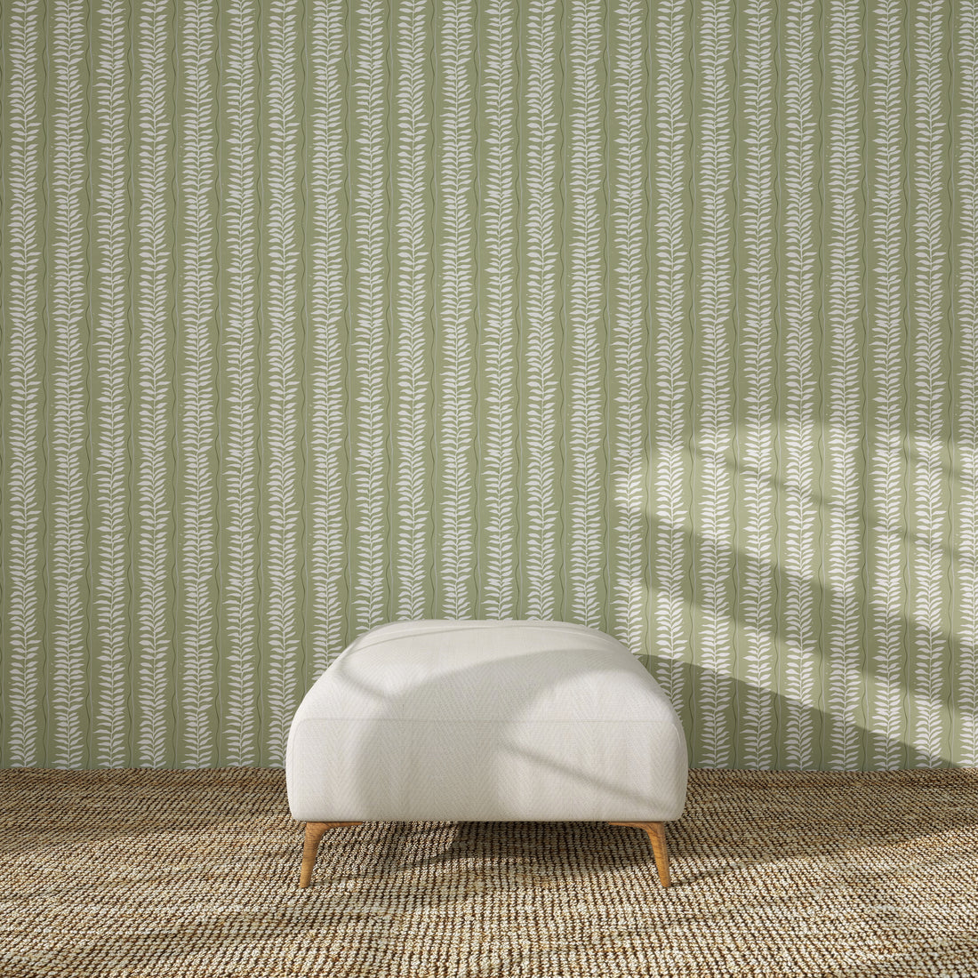 A white upholstered ottoman with wooden legs rests on a textured brown rug, in front of a green wall decorated with the Meadow Leaf Stripe Wallpaper Sample in Sage. Sunlight casts soft shadows across the scene.