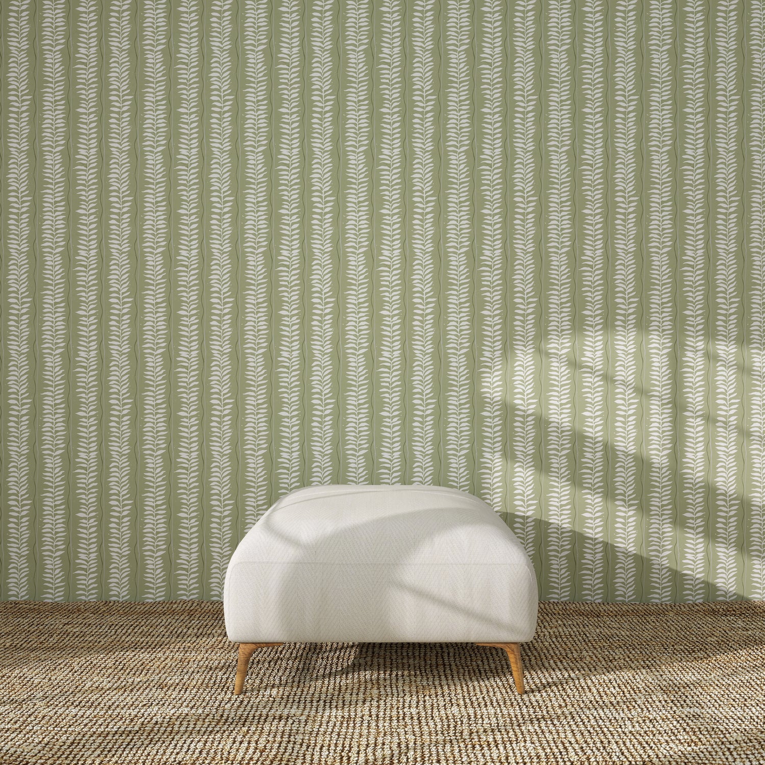 A white upholstered ottoman with wooden legs rests on a textured brown rug, in front of a green wall decorated with the Meadow Leaf Stripe Wallpaper Sample in Sage. Sunlight casts soft shadows across the scene.
