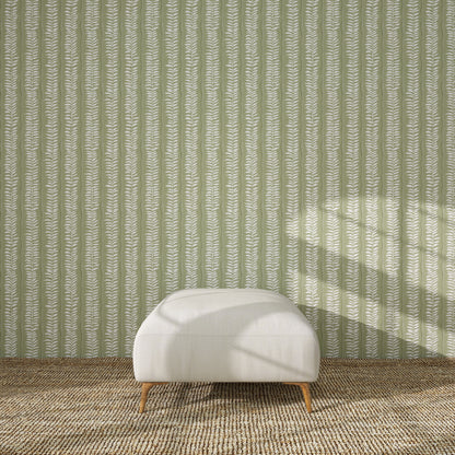 A white upholstered ottoman with wooden legs rests on a textured brown rug, in front of a green wall decorated with the Meadow Leaf Stripe Wallpaper Sample in Sage. Sunlight casts soft shadows across the scene.