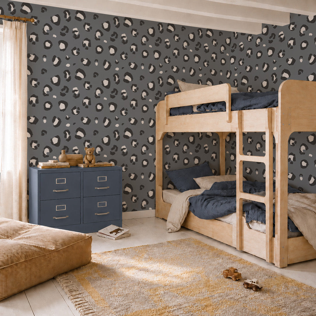 A cozy kids’ bedroom with light wood bunk beds, blue bedding, a blue dresser, tan pouf, yellow rug, large window, and bold Leopard Print Wallpaper in blue.