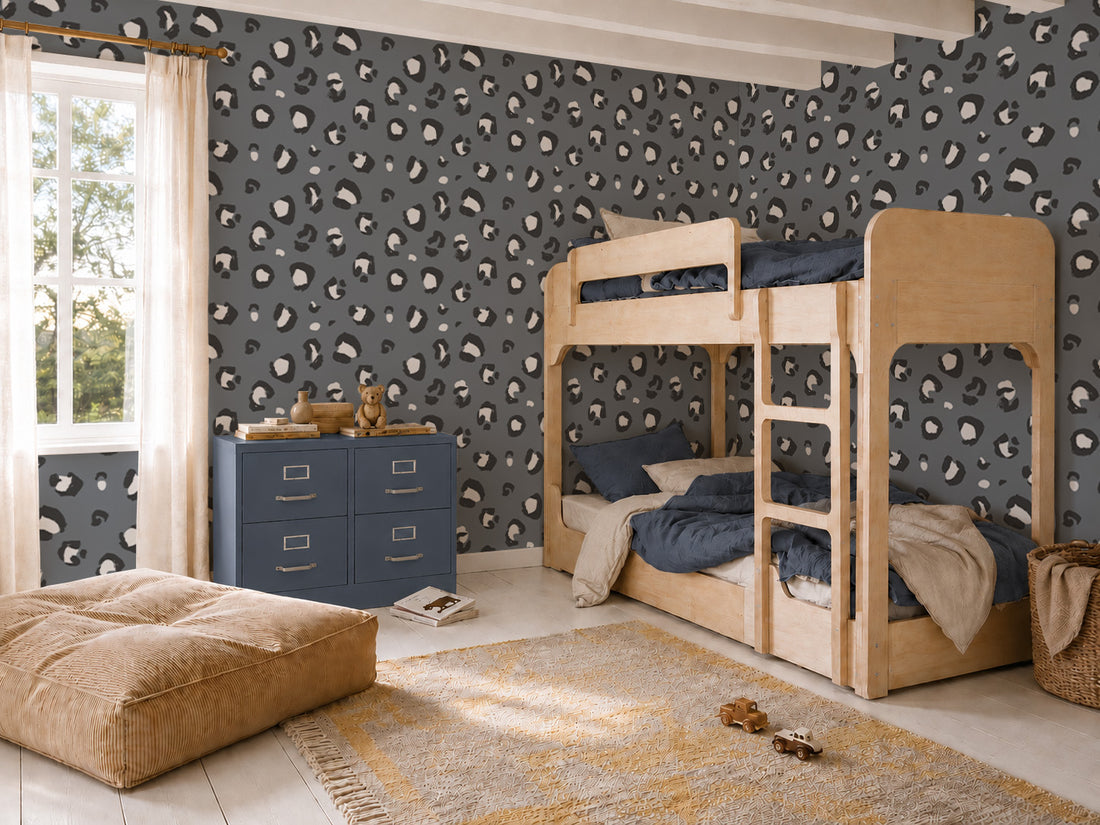 A cozy kids’ bedroom with light wood bunk beds, blue bedding, a blue dresser, tan pouf, yellow rug, large window, and bold Leopard Print Wallpaper in blue.