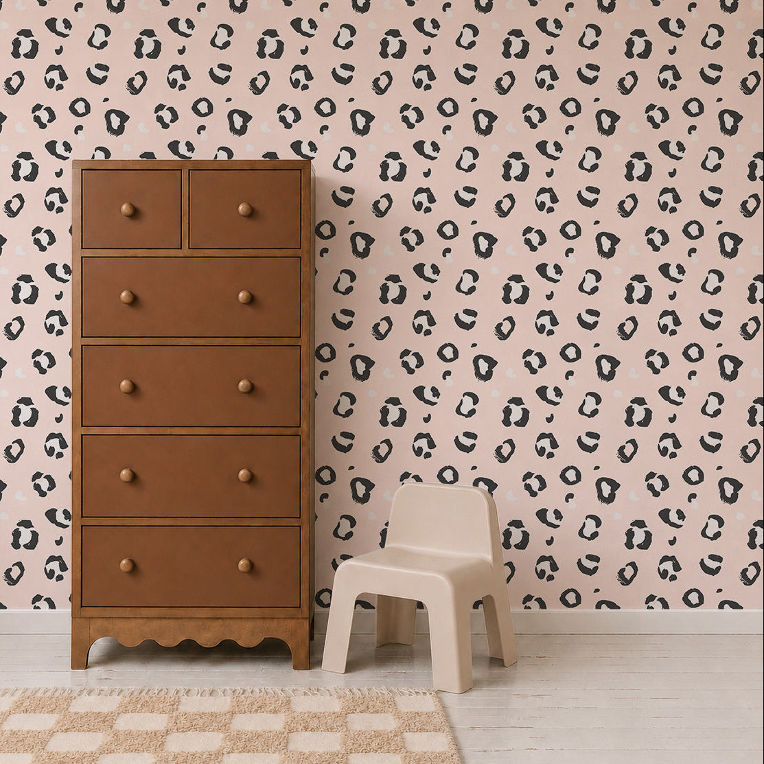 A brown dresser stands on a light rug next to a small beige chair and stool, set against a pink wall adorned with the Leopard Print Wallpaper, featuring a quirky black and white irregular dot pattern reminiscent of leopard spots.