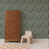 A tall brown dresser stands beside two stacked beige stools, set against a green wall covered in the Leopard Print Wallpaper, Sage, with a light wood floor, creating a striking statement in home decor.