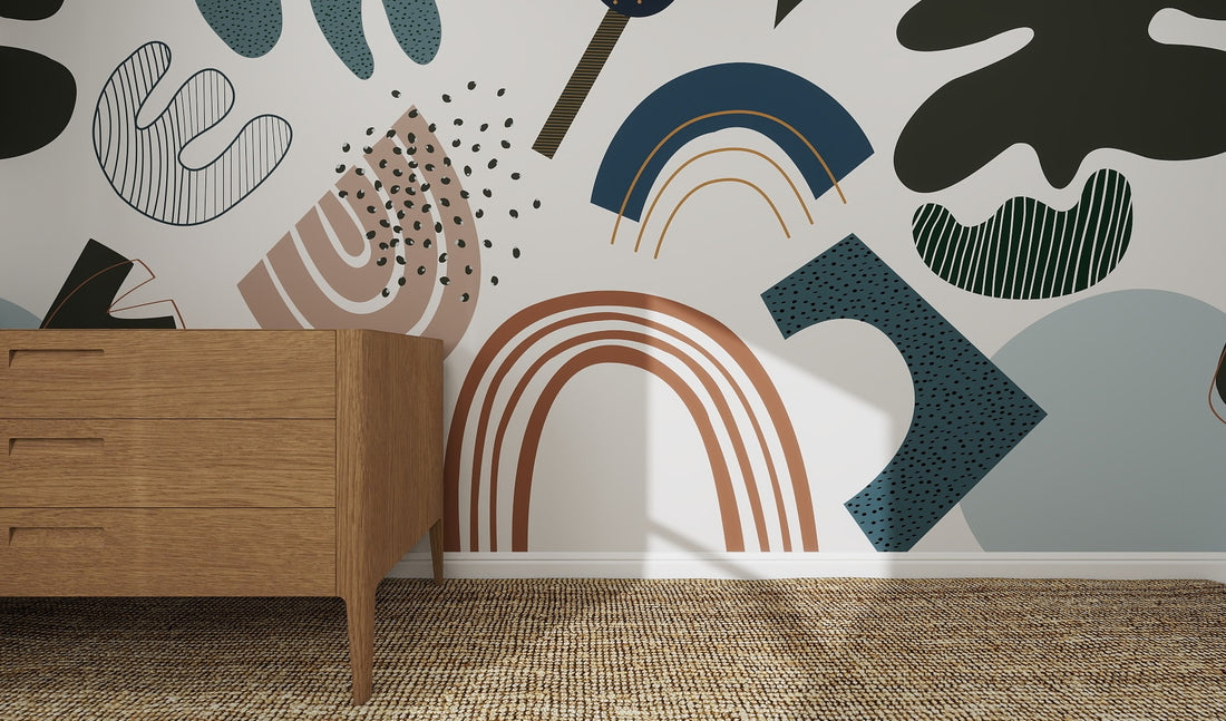 A wooden dresser stands on a woven rug by a white wall decorated with the Little Erin Abstract Wallpaper Mural | Blue, featuring bold muted shapes in blue, brown, green, and black for a modern artistic feel.