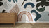 A wooden dresser stands on a woven rug by a white wall decorated with the Little Erin Abstract Wallpaper Mural | Blue, featuring bold muted shapes in blue, brown, green, and black for a modern artistic feel.