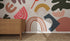 A wooden dresser stands on a woven rug beside a wall decorated with the Little Erin Abstract Wallpaper Mural, featuring colorful abstract shapes, rainbows, and vibrant pink, yellow, green, orange, and blue patterns.