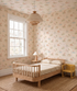 A cozy children’s bedroom with a wooden bed, checkered bedding, a side table and lamp, a yellow chair, and whimsical Little Friends Day Out Wallpaper—playful illustrations brighten the space filled with natural light from a large window.