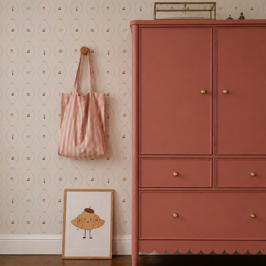 A pink dresser with gold knobs stands on a wooden floor beside a wall covered in Little Patisserie Stripe Wallpaper. A striped tote bag hangs nearby, and a framed smiling cupcake illustration leans against the wall.