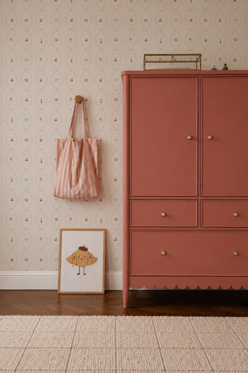 A pink dresser with gold knobs stands on a wooden floor beside a wall covered in Little Patisserie Stripe Wallpaper. A striped tote bag hangs nearby, and a framed smiling cupcake illustration leans against the wall.