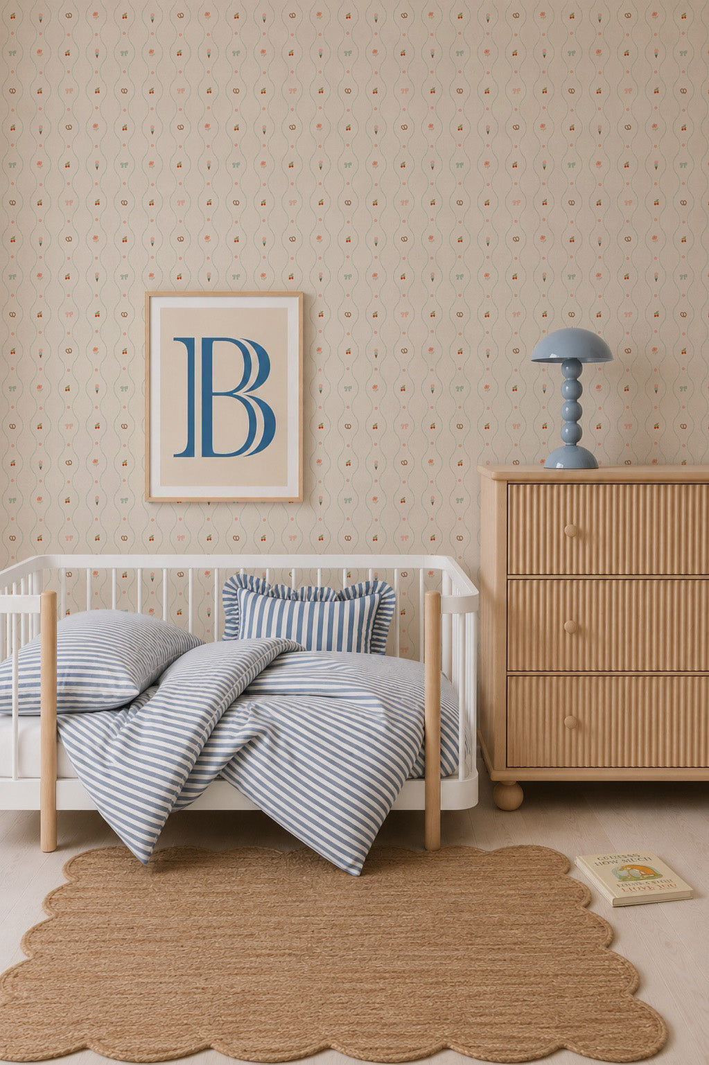 A cozy nursery features a small crib with blue striped bedding, a wooden dresser with a blue lamp, a scalloped rug, and a framed B on charming Little Patisserie Stripe Wallpaper. A children&