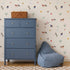 A blue chest of drawers and a bean bag chair sit in front of Little Paddock Wallpaper, which features a repeating pattern of horses, small houses, stars, and flowers.