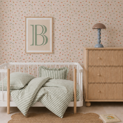 A nursery with a wooden crib, green striped bedding, a rattan dresser topped with a lamp and three stacked books, a rug, and Lucys Spring Buds Wallpaper featuring a delicate repeat pattern. A framed letter B hangs above the crib.
