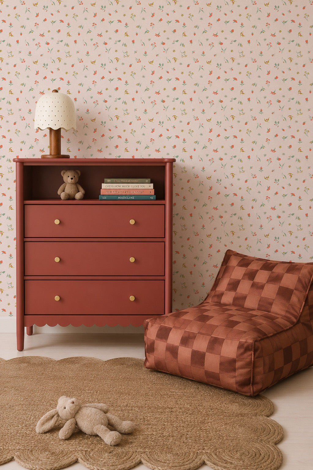 A children’s room with a red dresser, books, a teddy bear, a polka-dot lamp, a woven round rug, a plush bunny, and a brown checkered lounge chair is set against charming floral wallpaper featuring the delightful repeat pattern of Lucy’s Spring Buds Wallpaper.