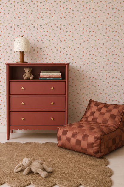 A children’s room with a red dresser, books, a teddy bear, a polka-dot lamp, a woven round rug, a plush bunny, and a brown checkered lounge chair is set against charming floral wallpaper featuring the delightful repeat pattern of Lucy’s Spring Buds Wallpaper.