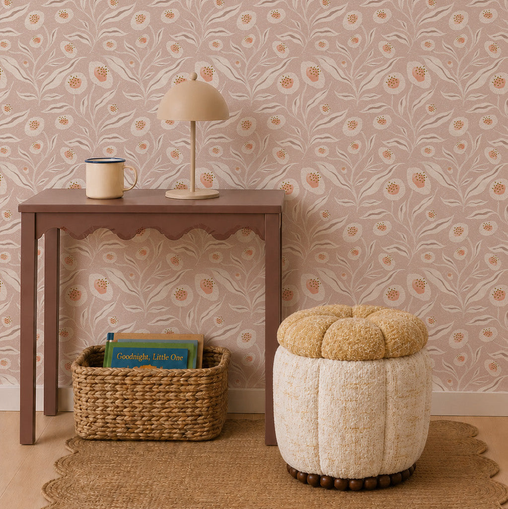 A small wooden table with a lamp and container sits against Matildas Sunflower Wallpaper in Neutral. A woven basket filled with books and a round textured ottoman are placed atop a natural fiber rug.