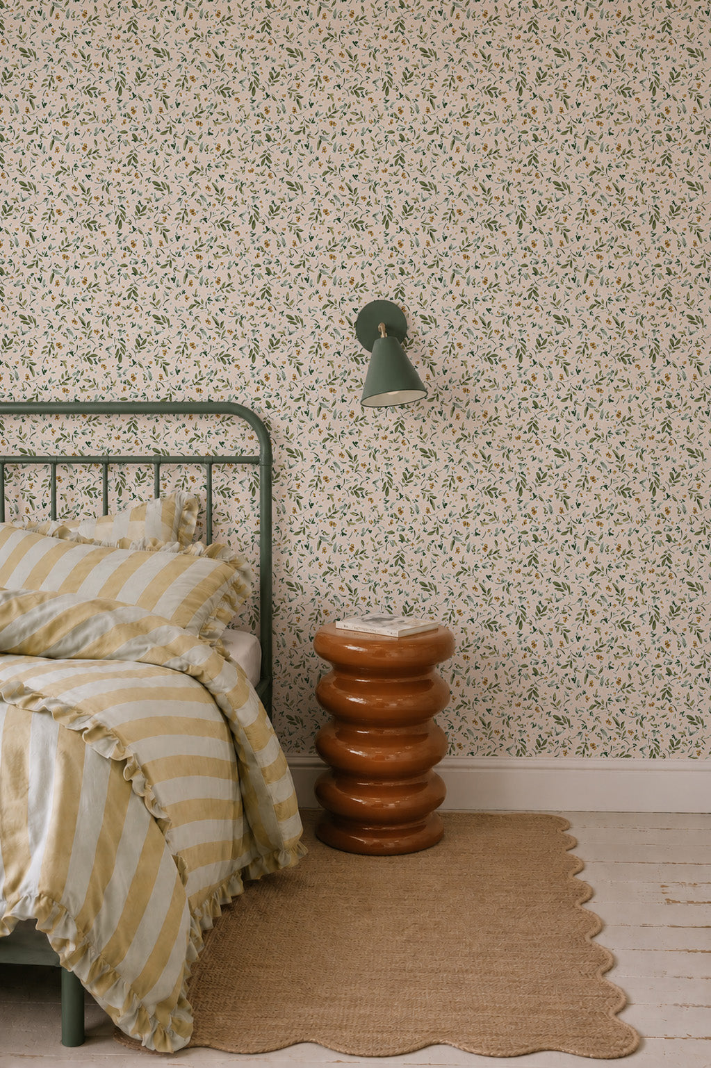 A cozy bedroom with a green metal bed, yellow and white striped bedding, a brown sculptural side table, a tan woven rug, and Meadow Flutter Leaves Wallpaper with hand-painted leaves, plus a green wall lamp above the table.