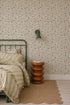 A cozy bedroom with a green metal bed, yellow and white striped bedding, a brown sculptural side table, a tan woven rug, and Meadow Flutter Leaves Wallpaper with hand-painted leaves, plus a green wall lamp above the table.