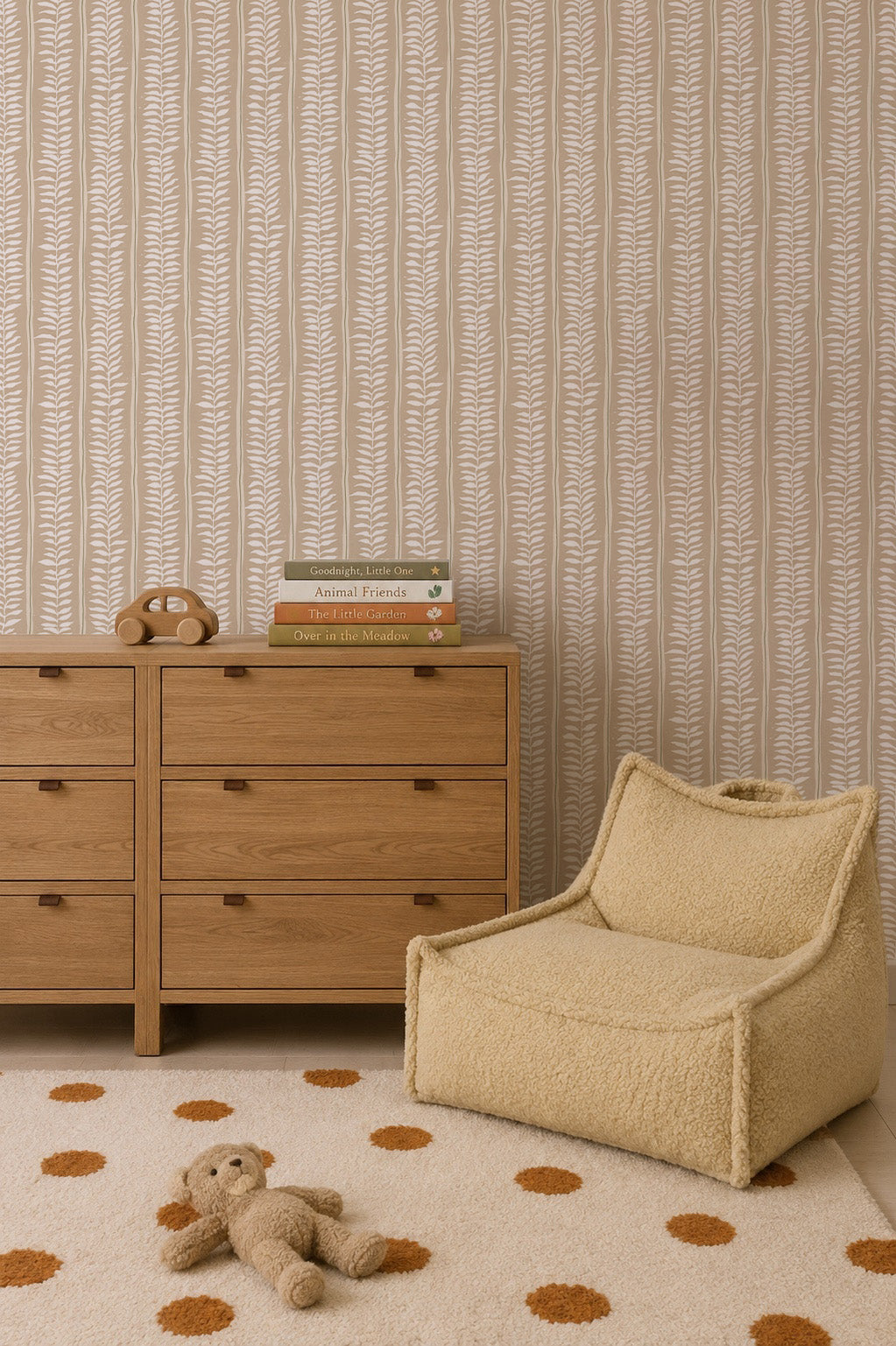A cozy kids’ room features a wooden dresser, children’s books, a toy phone, a beige textured chair, and a white rug with orange polka dots. A plush bear rests on the rug. Meadow Leaf Stripe Wallpaper, Clay brings subtle leafy charm to the space.
