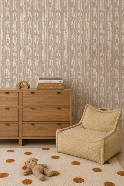 A cozy kids’ room features a wooden dresser, children’s books, a toy phone, a beige textured chair, and a white rug with orange polka dots. A plush bear rests on the rug. Meadow Leaf Stripe Wallpaper, Clay brings subtle leafy charm to the space.