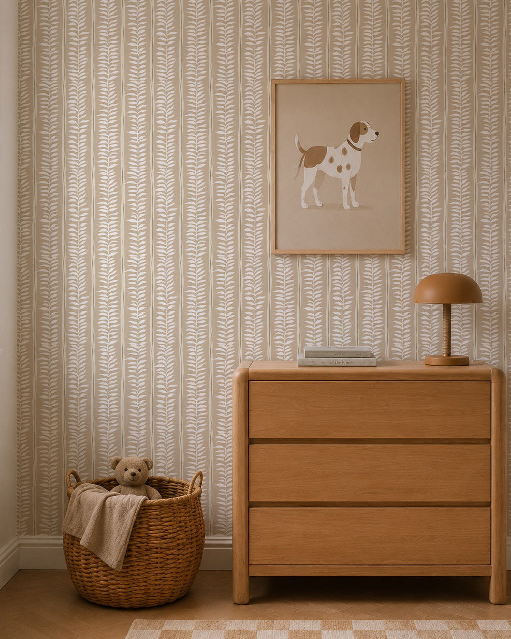A cozy room features a light wood dresser, brown lamp, books, and a framed dog illustration above. By the dresser sits a woven basket with a teddy bear and blanket. The wall is adorned with Meadow Leaf Stripe Wallpaper in Clay.