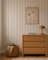 A cozy room features a light wood dresser, brown lamp, books, and a framed dog illustration above. By the dresser sits a woven basket with a teddy bear and blanket. The wall is adorned with Meadow Leaf Stripe Wallpaper in Clay.
