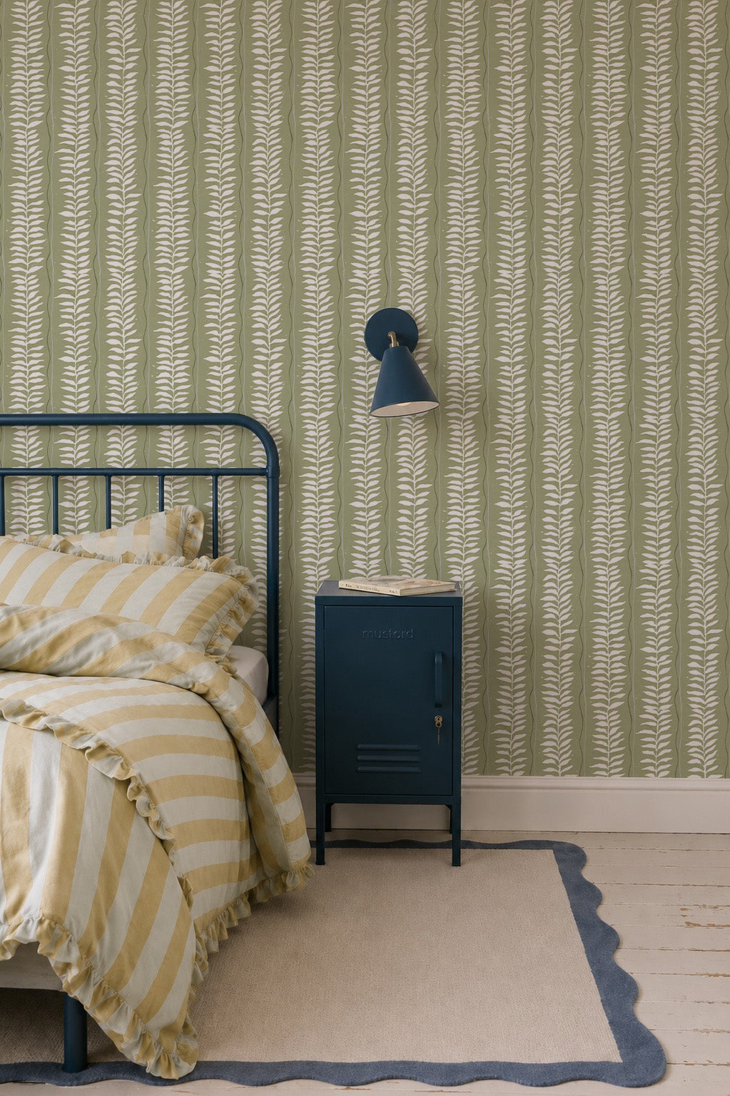 A cozy bedroom with a metal bed frame, yellow botanical stripe bedding, a blue nightstand, a wall-mounted blue lamp, Meadow Leaf Stripe Wallpaper in Sage, and a beige rug with a blue border on the white wooden floor.