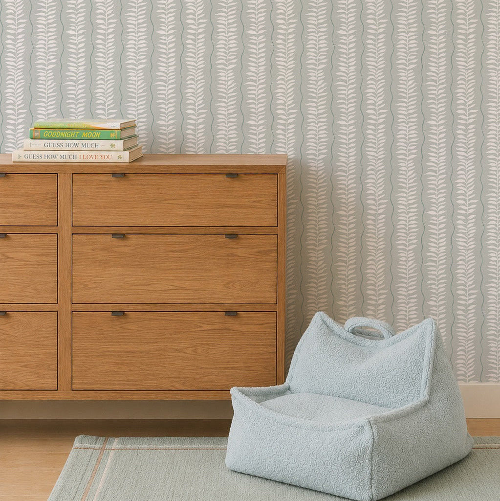 A wooden dresser with books sits beside a pale blue textured pet bed on a rug, set against Meadow Leaf Stripe Wallpaper in Sea Foam with vertical leafy stripes in the background.