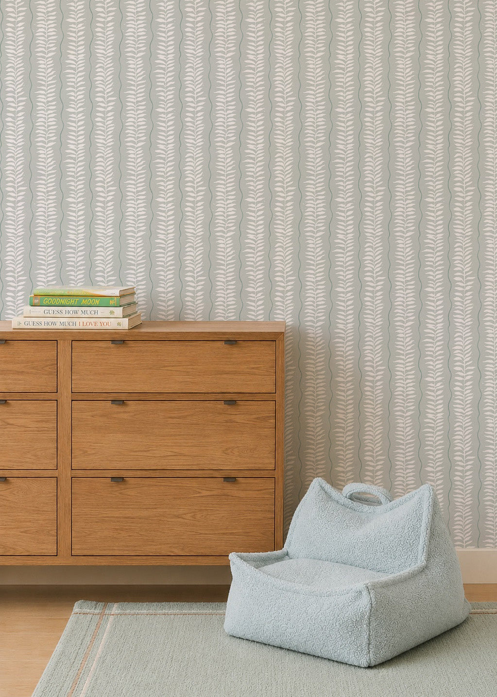A wooden dresser with books sits beside a pale blue textured pet bed on a rug, set against Meadow Leaf Stripe Wallpaper in Sea Foam with vertical leafy stripes in the background.