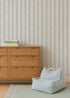 A wooden dresser with books sits beside a pale blue textured pet bed on a rug, set against Meadow Leaf Stripe Wallpaper in Sea Foam with vertical leafy stripes in the background.