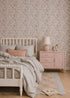 A cozy bedroom features a light wood bed, soft gray bedding, and pastel decor. Millie Floral Wallpaper and a pink nightstand add gentle charm, while a toy giraffe rests beneath the warm glow of a pink lamp.