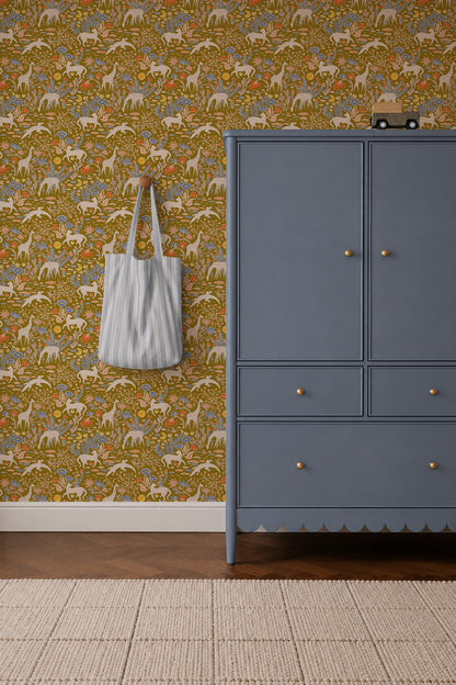 A blue dresser with gold knobs sits on a beige rug near a wall accented by Mils Menagerie Wallpaper, Gold, featuring yellow, orange, and white dinosaurs. A blue and white striped tote bag hangs from a hook nearby.