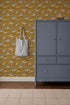 A blue dresser with gold knobs sits on a beige rug near a wall accented by Mils Menagerie Wallpaper, Gold, featuring yellow, orange, and white dinosaurs. A blue and white striped tote bag hangs from a hook nearby.