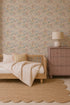 A cozy bedroom featuring a wooden daybed, striped pillows and blanket, a scalloped beige rug, a mauve dresser with a lamp, and elegant Mils Menagerie Wallpaper in Rose with soft pastel floral patterns.