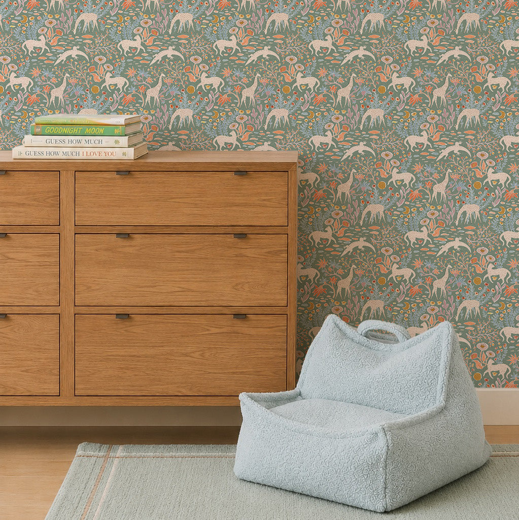 A wooden dresser with books stands by a pale blue chair on a green rug. The wall is adorned with Mils Menagerie Wallpaper, Sage, featuring white horses and orange flowers on a green background.