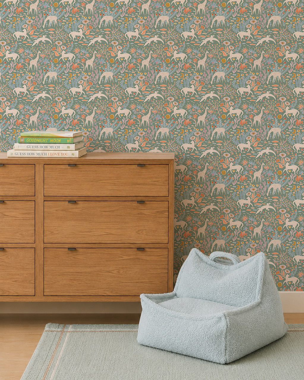 A wooden dresser with books stands by a pale blue chair on a green rug. The wall is adorned with Mils Menagerie Wallpaper, Sage, featuring white horses and orange flowers on a green background.