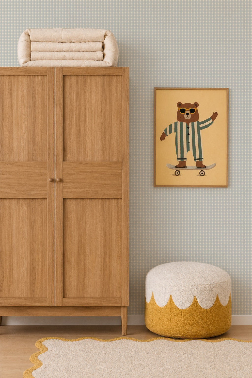 A wooden wardrobe stands next to a round pouf and a beige rug. Above the pouf is a framed picture of a bear in striped pajamas riding a skateboard, set against charming Mini Check Wallpaper in Blue.