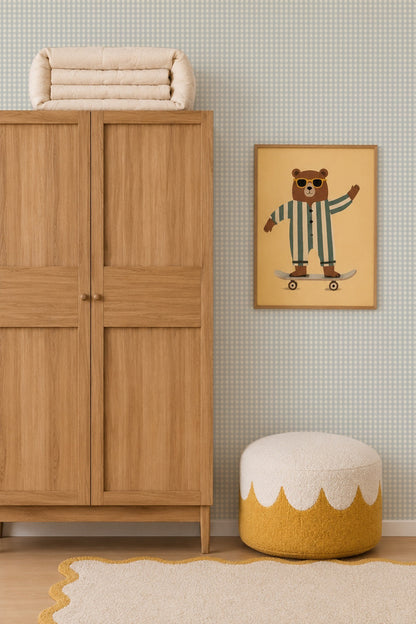 A wooden wardrobe stands next to a round pouf and a beige rug. Above the pouf is a framed picture of a bear in striped pajamas riding a skateboard, set against charming Mini Check Wallpaper in Blue.