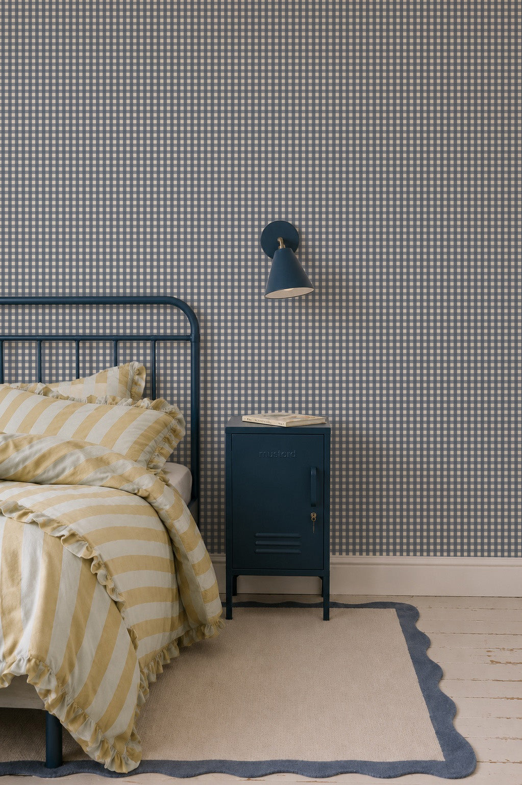 A dark blue metal locker-style bedside table sits next to a bed with yellow striped bedding against Mini Check Wallpaper, Deep Blue.