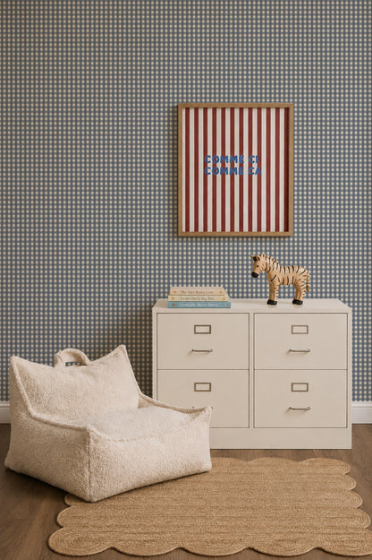 A cozy corner featuring a cream-colored lounge chair, a small metal filing cabinet with a wooden zebra figurine and books on top, and a framed red and white striped print hanging on Mini Check Wallpaper, Deep Blue.
