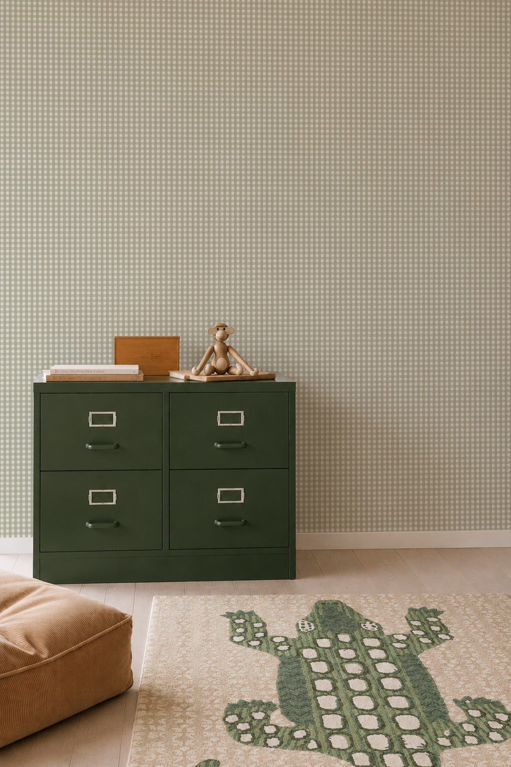 A green four-drawer metal cabinet stands against a wall with Mini Check Wallpaper, Green, topped with a wooden monkey figurine and a wooden box, with a green crocodile-shaped rug on the floor.