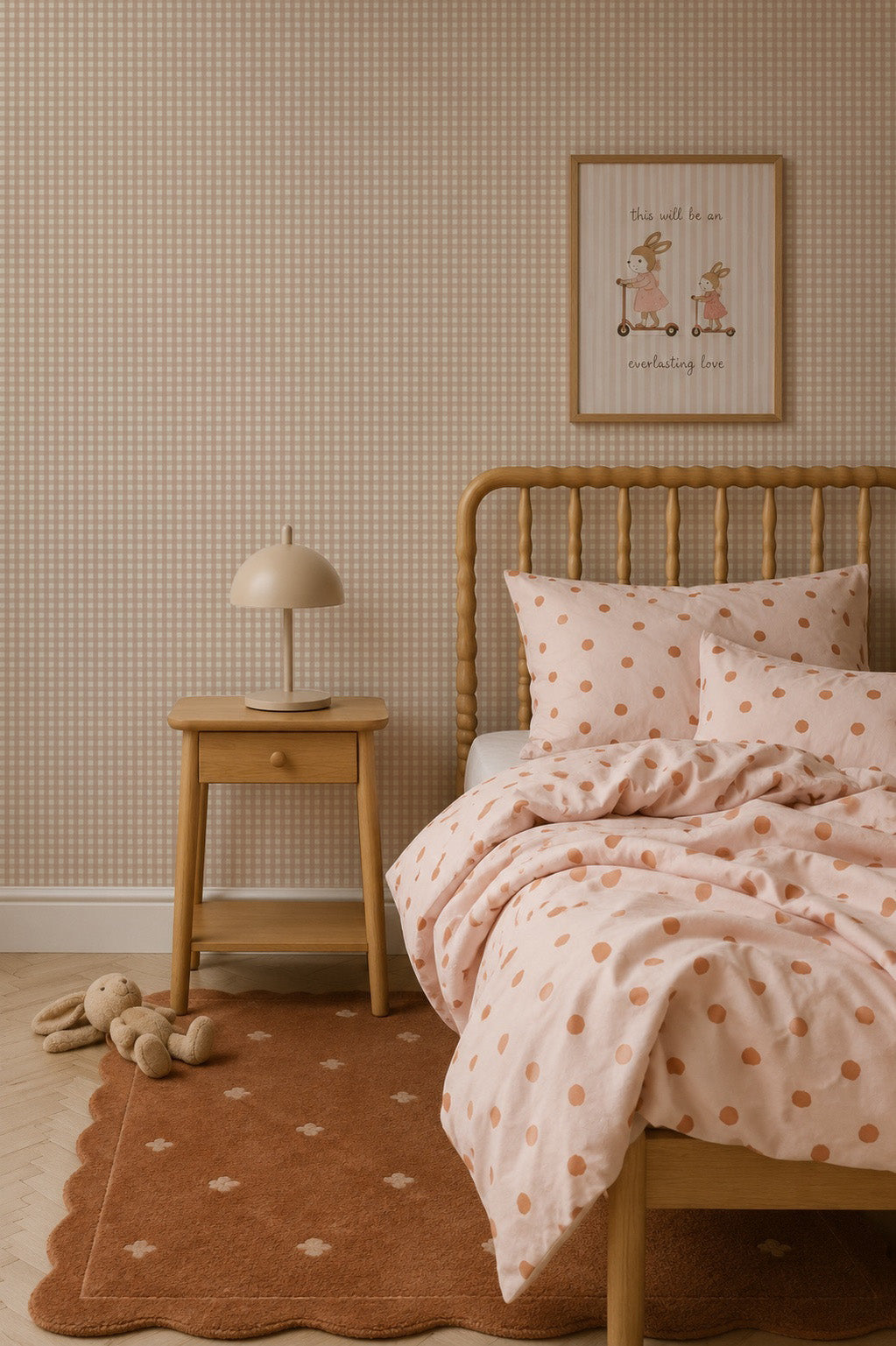 A cozy bedroom features a wooden bed with pink polka dot bedding, a matching nightstand with a beige lamp, a stuffed animal on a brown rug, and framed art on walls adorned with Mini Check Wallpaper, Pink—a charming choice perfect for adding a playful touch reminiscent of classic children’s wallpaper.