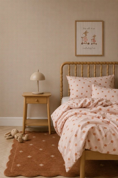 A cozy bedroom features a wooden bed with pink polka dot bedding, a matching nightstand with a beige lamp, a stuffed animal on a brown rug, and framed art on walls adorned with Mini Check Wallpaper, Pink—a charming choice perfect for adding a playful touch reminiscent of classic children’s wallpaper.