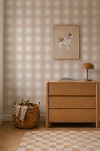A cozy nursery features a wicker basket with a teddy bear, a wooden dresser with books and a lamp, and a framed dog illustration hanging above against the charming Mini Check Wallpaper in Pink.