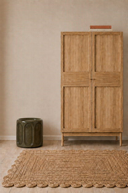A minimalist room with a wooden wardrobe, a textured tan rug, a green ceramic stool, and a book placed atop the wardrobe. The wall is adorned with Mini Check Wallpaper, Pink—a subtly patterned wallpaper ideal for children’s rooms.