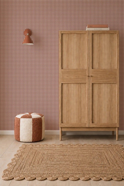 A wooden wardrobe stands against Mini Check Wallpaper, Red, with a patterned pouf, wall sconce, and textured rug in the foreground.