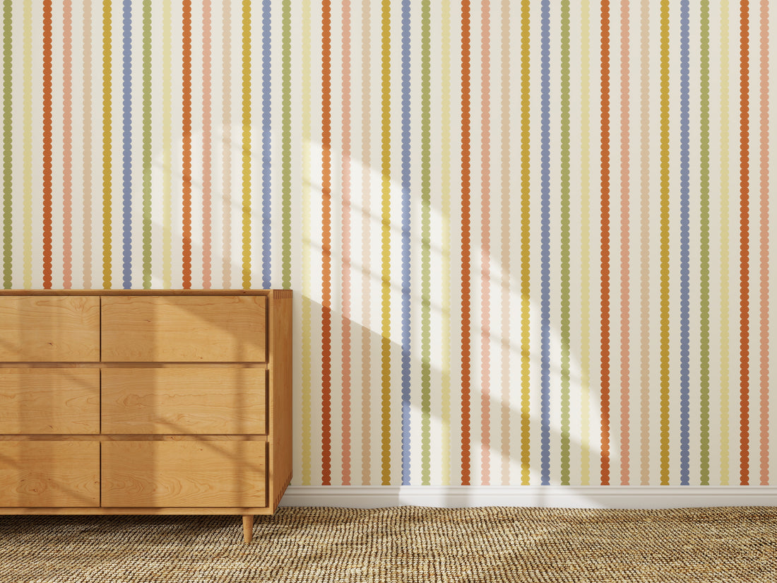 A wooden dresser stands on a beige carpet beside a wall decorated with the Mini Scallop Stripe Wallpaper Repeat Pattern | Multi. Sunlight casts window shadows across the wall and part of the dresser.