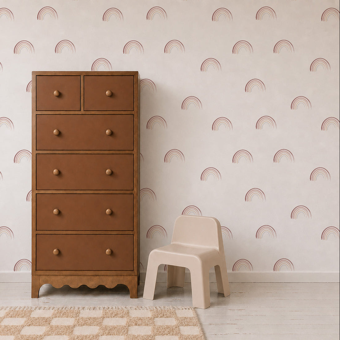 A tan dresser and a small beige chair sit on a white floor with a tan-checkered rug. The wall behind them is decorated with Mini Watercolour Rainbow Wallpaper, featuring a charming pink rainbow pattern that adds a playful and artistic touch to the room.