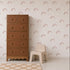 A tan dresser and a small beige chair sit on a white floor with a tan-checkered rug. The wall behind them is decorated with Mini Watercolour Rainbow Wallpaper, featuring a charming pink rainbow pattern that adds a playful and artistic touch to the room.