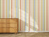 A wooden dresser stands on a beige carpet before a wall adorned with Mini Scallop Stripe Wallpaper Repeat Pattern Multi, its vibrant orange, green, yellow, and blue stripes showcased beautifully as sunlight casts gentle shadows to enhance the effect.