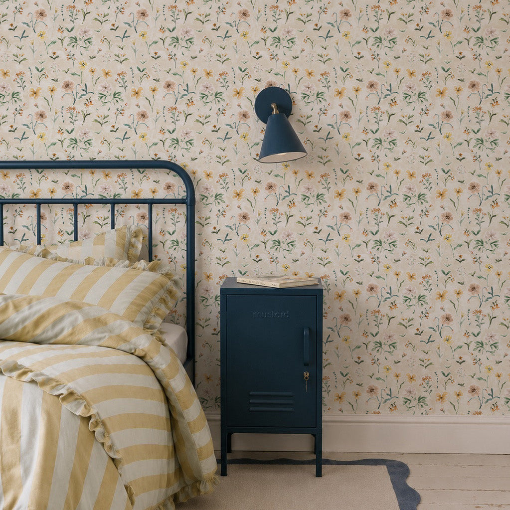 Cozy bedroom corner with a metal bed, striped yellow bedding, a blue nightstand, and a blue wall lamp. The wall is decorated with Morning Meadow Wallpaper, bringing a delightful touch of hand-painted wildflowers that’s perfect for kids’ spaces.